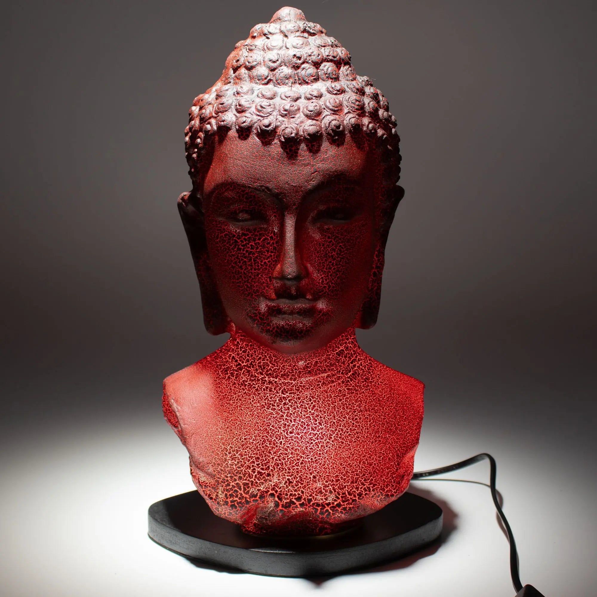 A glowing Buddha bust lamp with a textured red finish on an oval black stand, featuring intricate detailing and serene facial expression, Buddha Bust Lamp Molton Lava - Oval stand.