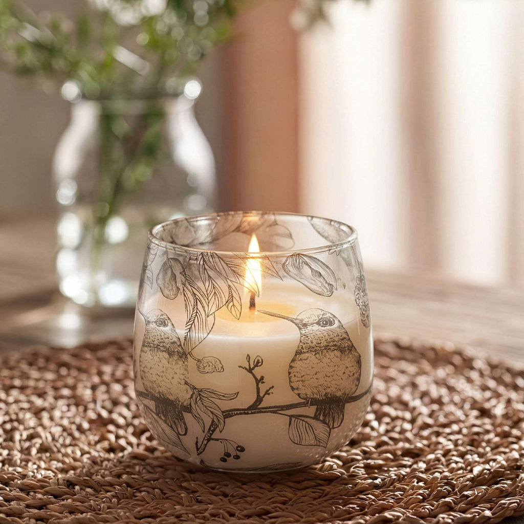 Medium Glass Jar Candles - Drawn by Flame featuring a lit candle in a glass jar adorned with delicate black bird and leaf illustrations, placed on a textured woven mat in soft natural light.