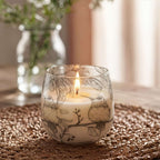 Medium Glass Jar Candles - Drawn by Flame featuring a lit candle in a glass jar adorned with delicate black bird and leaf illustrations, placed on a textured woven mat in soft natural light.