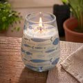 Tall Glass Jar Candles - Seabreeze featuring a lit candle in a clear glass jar decorated with blue fish patterns, placed on a wooden surface near a beige cloth.