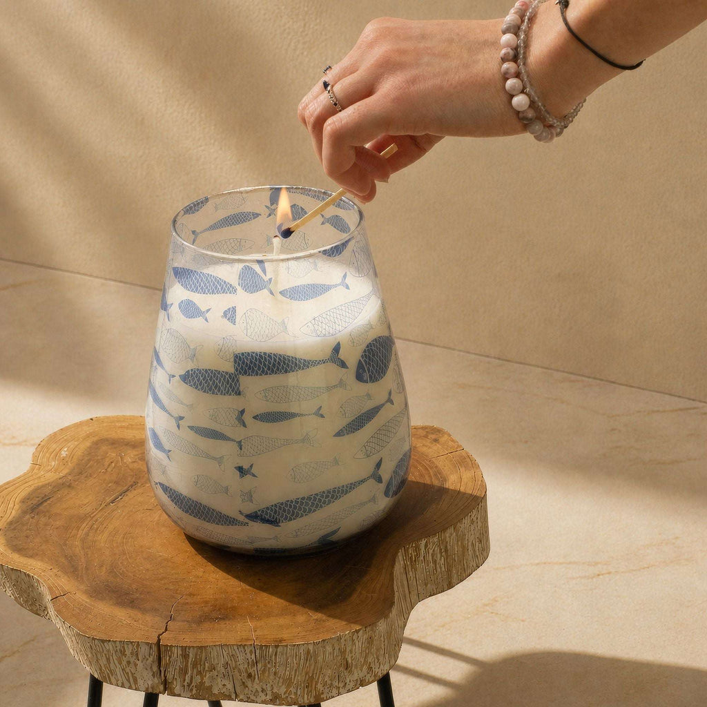 Hand lighting a Tall Glass Jar Candles - Seabreeze with blue fish pattern on a wooden surface in soft natural light.