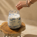 Hand lighting a Tall Glass Jar Candles - Seabreeze with blue fish pattern on a wooden surface in soft natural light.