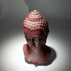 Buddha Bust Lamp Molton Lava - Oval stand featuring a textured Buddha head sculpture with a dark reddish lava-like finish, casting a serene shadow on a grey background.