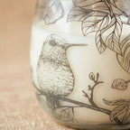 Close-up of a Medium Glass Jar Candles - Drawn by Flame featuring detailed black line art of a hummingbird perched on a branch surrounded by leaves on a white candle surface.
