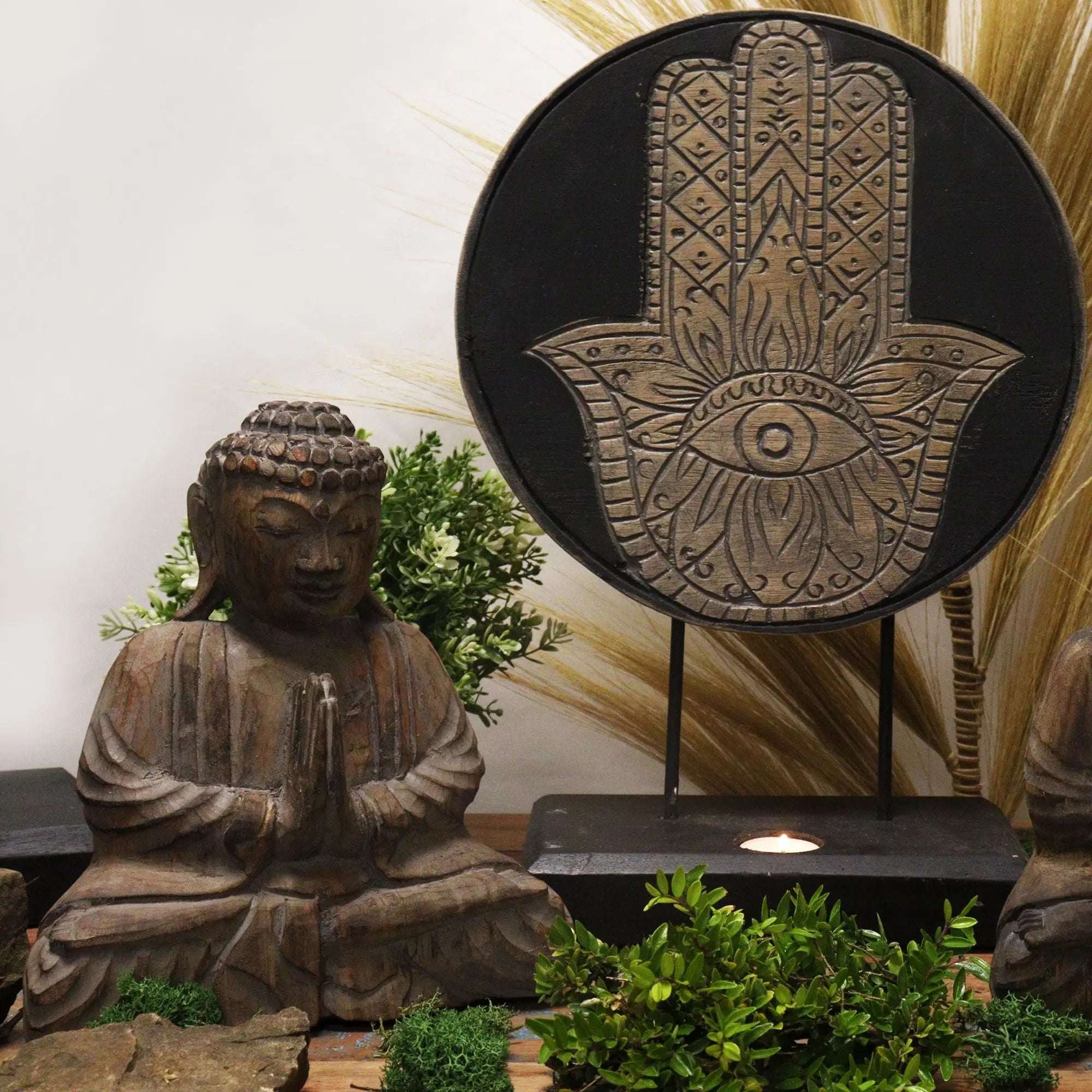 Buddha Feng Shui Set - Hamsa - Grey featuring a hand-carved Buddha statue in a meditative pose beside an artistic Hamsa wall hanging, styled with green foliage.