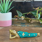 Brass Buddha Figure - Lying Down - 10cm with turquoise mosaic inlay, resting on a wooden surface next to a potted plant and trailing greenery, ideal for Buddha decor and tabletop display.