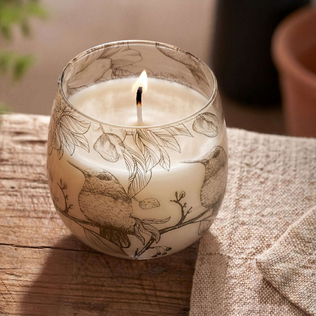 A lit Medium Glass Jar Candles - Drawn by Flame featuring delicate illustrations of birds and botanical elements on clear glass, placed on a wooden surface.