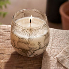 A lit Medium Glass Jar Candles - Drawn by Flame featuring delicate illustrations of birds and botanical elements on clear glass, placed on a wooden surface.