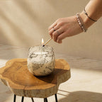 A hand with delicate rings lighting a fire with a matchstick in a Medium Glass Jar Candles - Drawn by Flame, placed on a rustic wooden table in natural light.