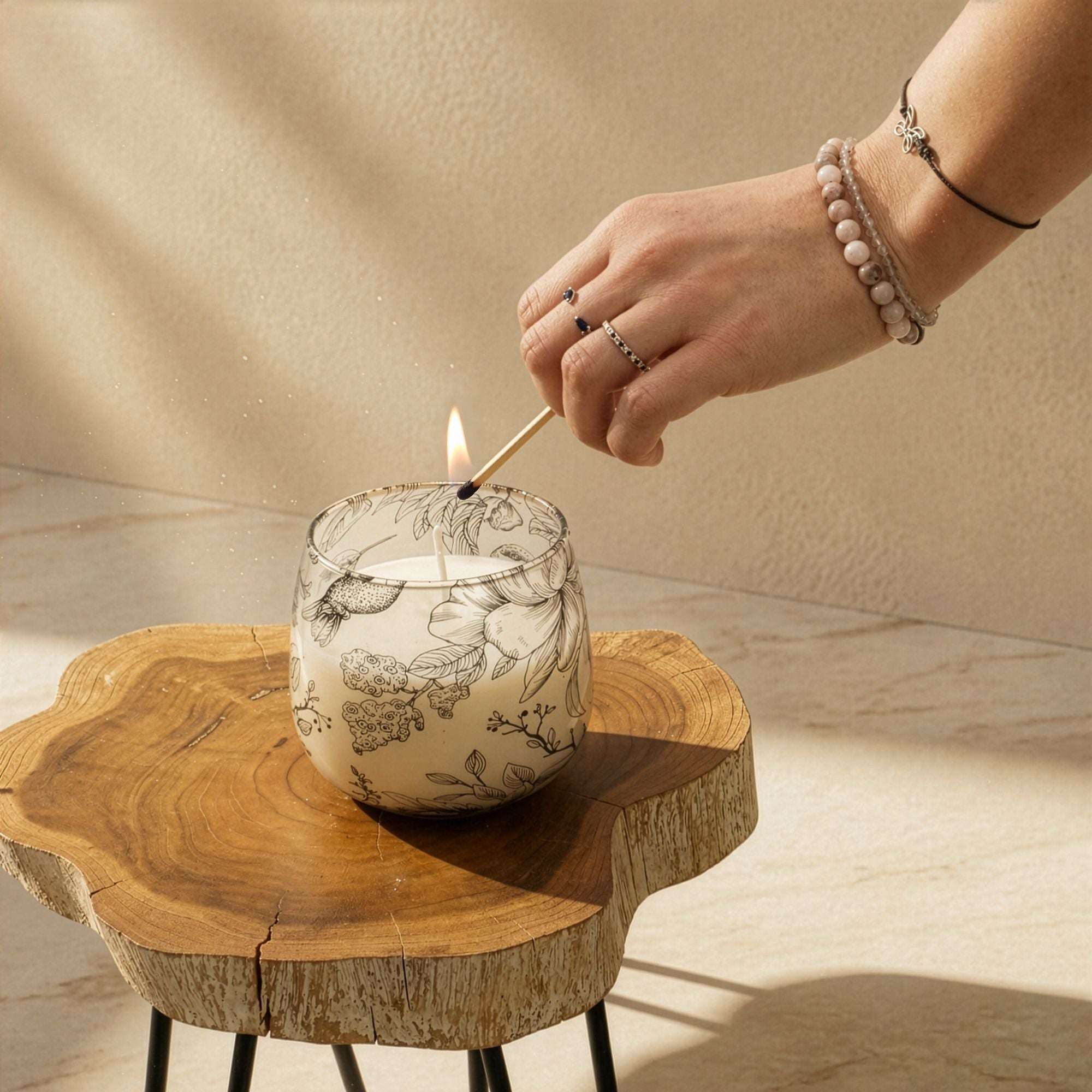 A hand with delicate rings lighting a fire with a matchstick in a Medium Glass Jar Candles - Drawn by Flame, placed on a rustic wooden table in natural light.