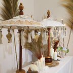 Two Bali Home Decor Parasol - Canvas - Gold Decor- 40cm with wooden handles, hanging gold and silver charms, white tassels, displayed on a table with candles and decorative dried plants.