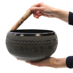 A hand holding a wooden striker rubbing the edge of a black bowl engraved with intricate designs, showcasing the Extra Loud - Singing Bowl - One Buddha.