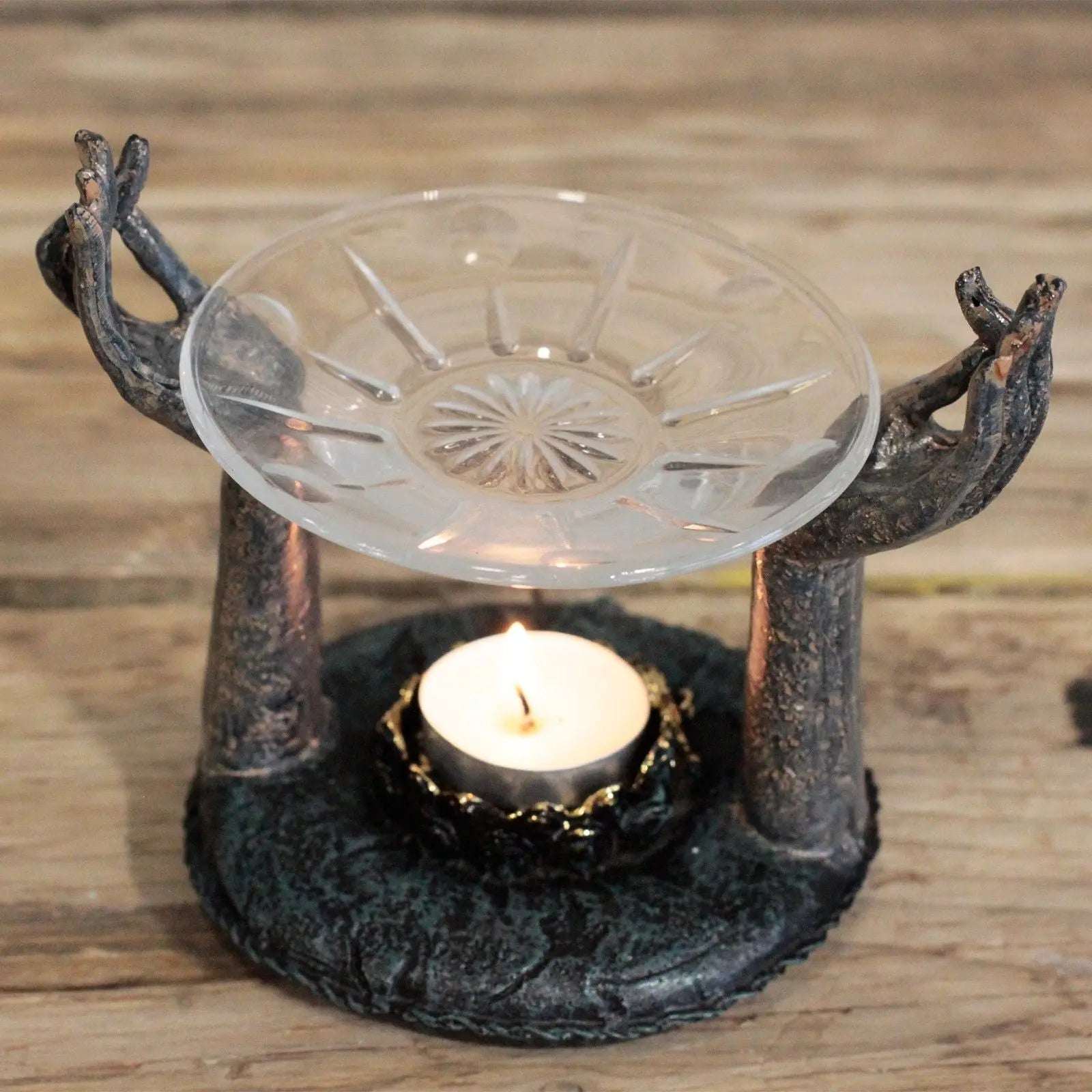 Antique Buddha - Om Hands Oil Burner with handcrafted metal hands holding a decorative glass dish above a lit tealight candle on a rustic wooden surface