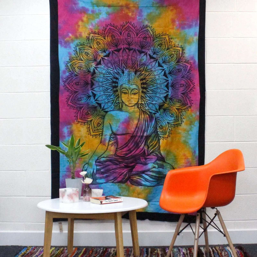 Colorful tie-dye fabric with a detailed Buddha design is displayed as a Single Cotton Bedspread + Wall Hanging - Peaceful Buddha hanging on a white wall above a modern table and orange chair.