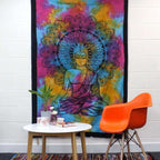 Colorful tie-dye fabric with a detailed Buddha design is displayed as a Single Cotton Bedspread + Wall Hanging - Peaceful Buddha hanging on a white wall above a modern table and orange chair.