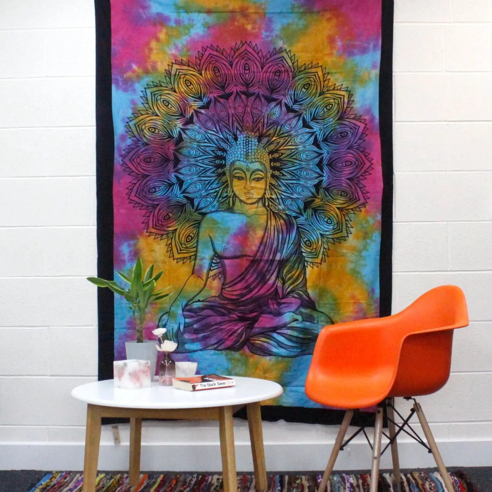 Colorful tie-dye wall hanging featuring a detailed Buddha figure with an intricate mandala halo behind, displayed above a round white table and orange chair, showcasing the Single Cotton Bedspread + Wall Hanging - Peaceful Buddha.