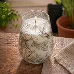 Tall Glass Jar Candles - Drawn by Flame featuring a lit candle in a glass jar decorated with delicate floral and hummingbird illustrations on a wooden surface.