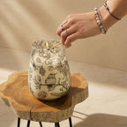 Hand lighting a Tall Glass Jar Candles - Drawn by Flame with intricate black botanical and bird illustrations on the glass, set on a natural wood slab table.