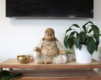 Happy Buddha - Whitewash 50cm statue in a serene seated pose on a wooden surface, surrounded by green plants and a brass bowl, creating a peaceful decorative setting.