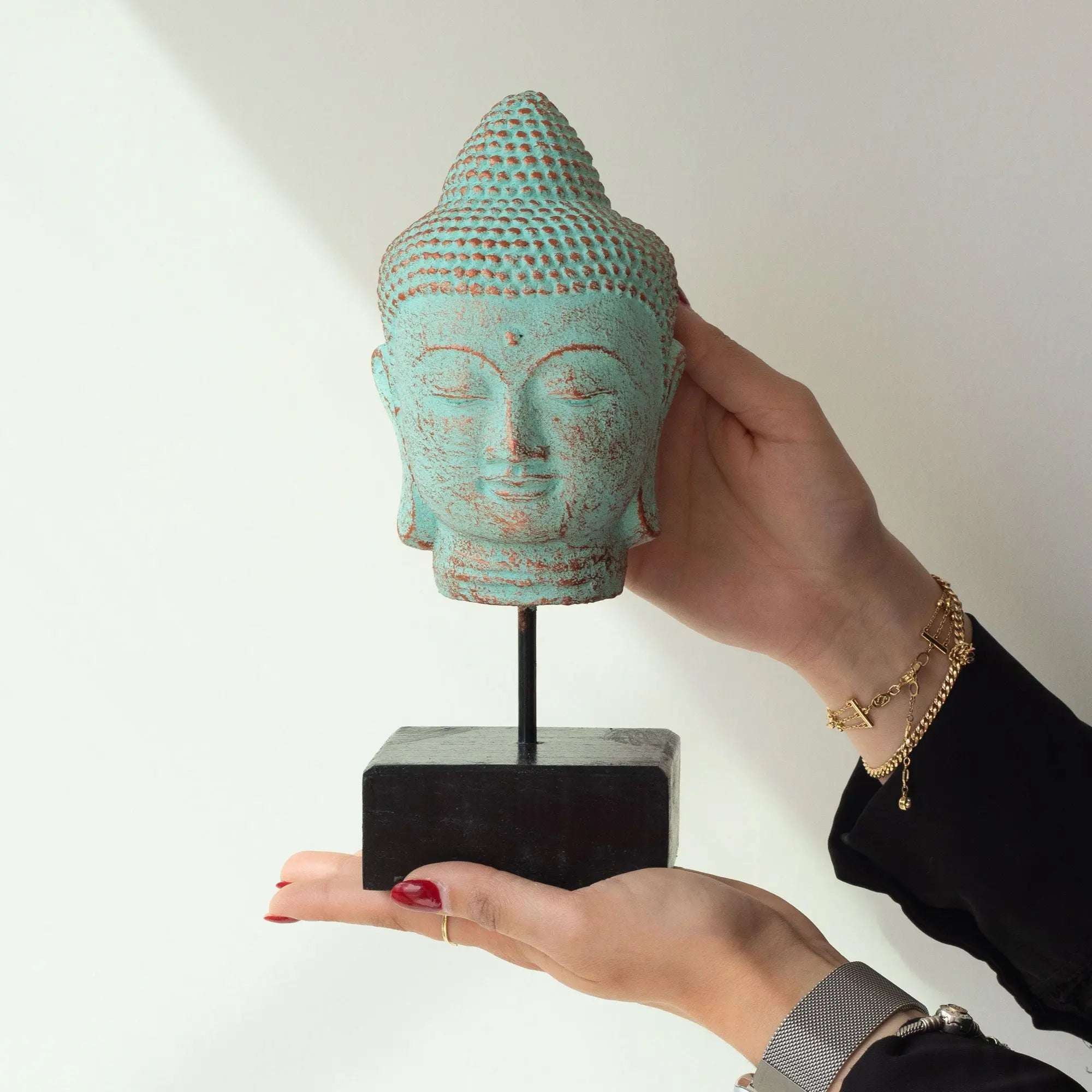Stillness Buddha Head on Stand - Green Copper displayed on a black stand, featuring a serene Buddha face with a textured green copper finish, held gently by two hands.
