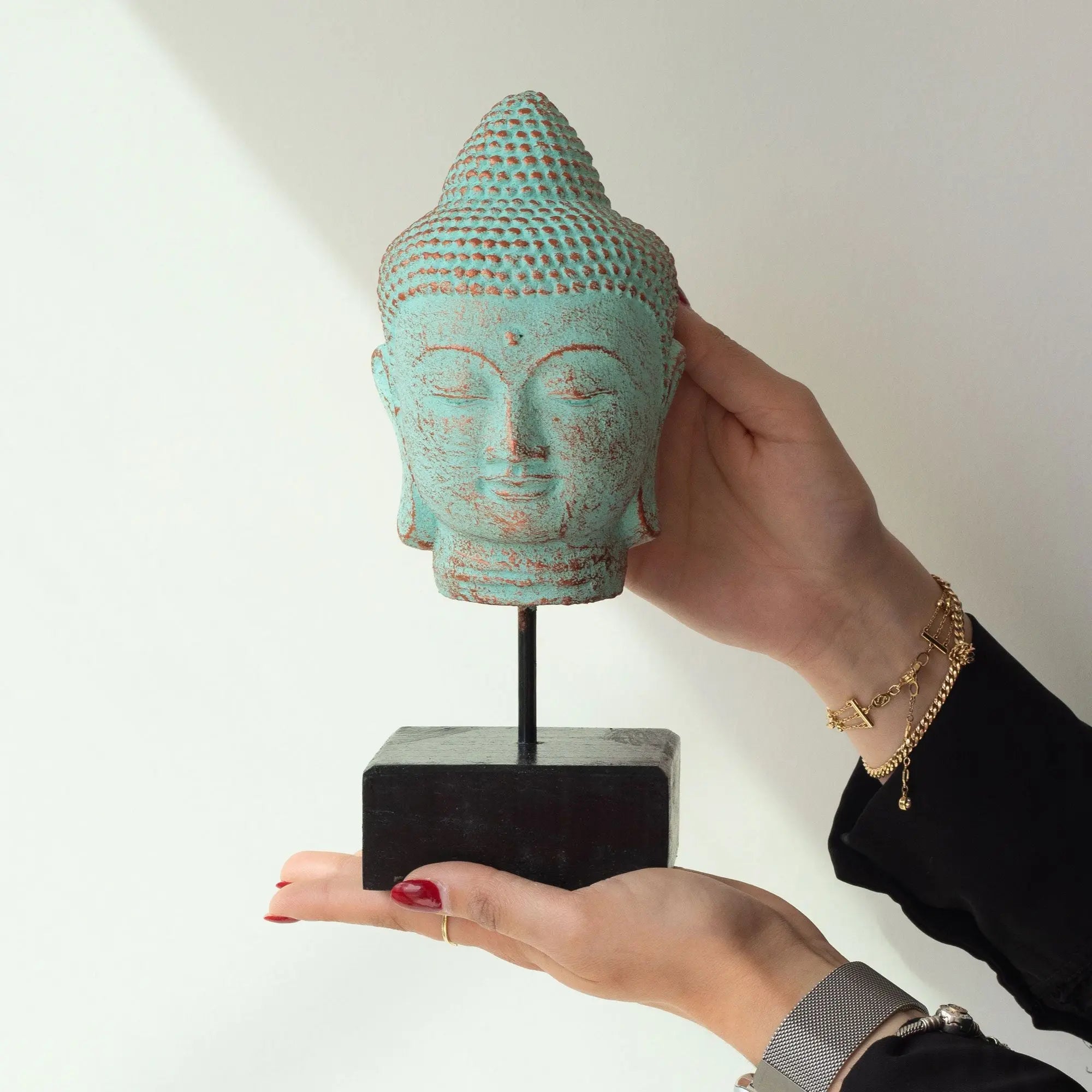 Stillness Buddha Head on Stand - Green Copper displayed on a black pedestal, held delicately by hands, featuring a serene-faced Buddha with textured green copper finish.