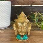Brass Buddha Figure - Lrg Head - 11.5 cm with intricate facial features and a turquoise inlay base, displayed on a wooden surface with green foliage and a white textured planter in the background.