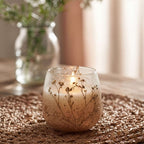 Small Glass Jar Candles - Wildlight Woods featuring a lit candle in a decorative glass jar with embedded dried floral accents, placed on a textured woven mat.