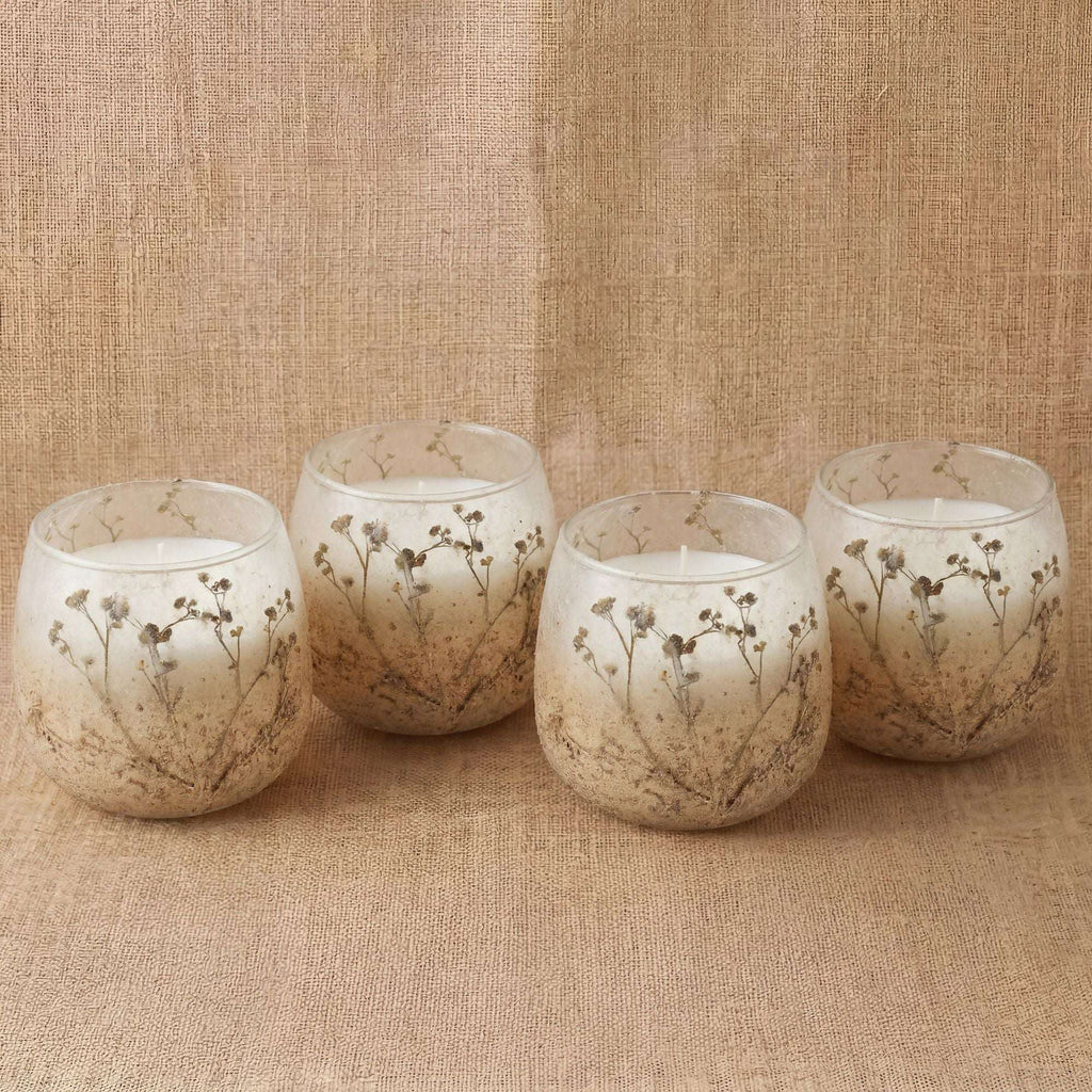A set of four Small Glass Jar Candles - Wildlight Woods featuring delicate dried floral designs encased in frosted glass, displayed on a textured beige fabric background.
