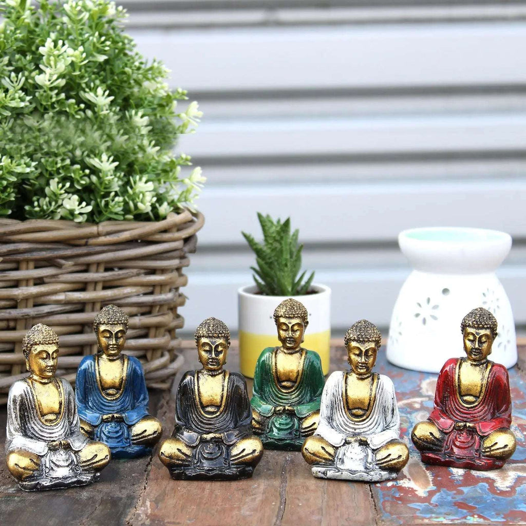 A collection of Gold Mini Buddha (Assorted Colours) statues in various seated meditation poses in blue, black, green, white, and red, displayed on a wooden surface with decorative plants in the background.