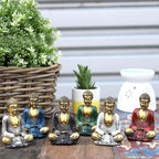 A collection of Gold Mini Buddha (Assorted Colours) statues in various seated meditation poses in blue, black, green, white, and red, displayed on a wooden surface with decorative plants in the background.