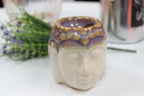 Close-up of a Buddha Oil Burner - Ivory & Mint with a serene Buddha face design, featuring earthy tones and detailed texture, set on a white surface with blurred background greenery.