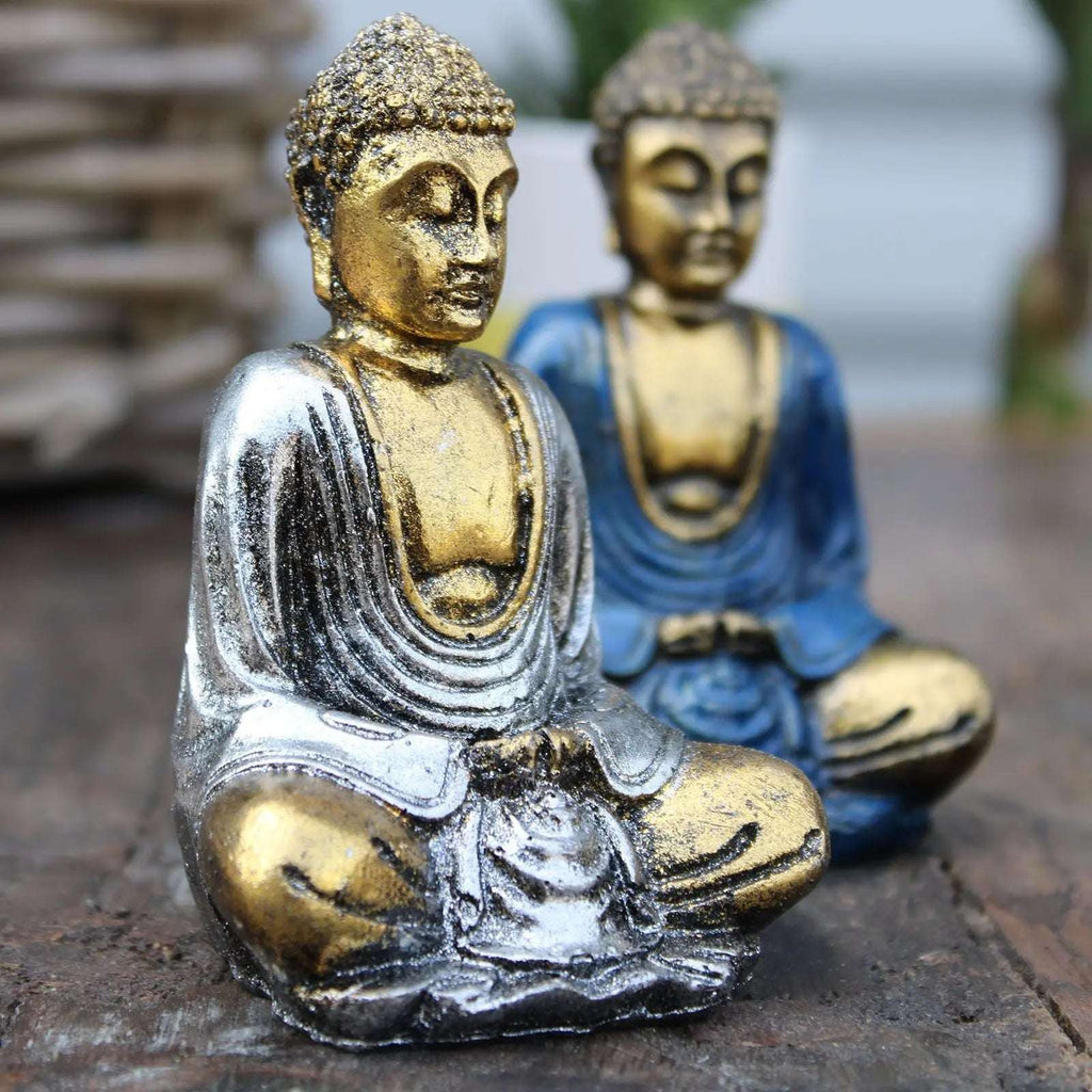 Two Gold Mini Buddha (Assorted Colours) statues in metallic silver and blue finishes seated in a meditative pose on a wooden surface with a blurred natural background.