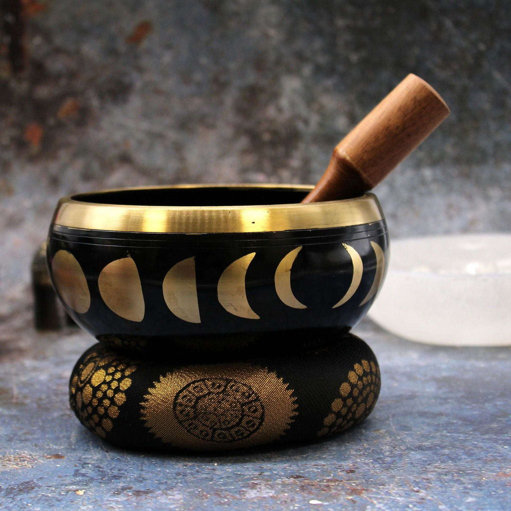 Close-up of a Large Moon Phase Singing Bowl Set- Black 14cm with moon phase designs in gold, sitting on a decorative cushion with a wooden mallet resting inside the bowl.