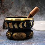 Close-up of a Large Moon Phase Singing Bowl Set- Black 14cm with moon phase designs in gold, sitting on a decorative cushion with a wooden mallet resting inside the bowl.