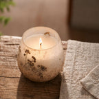 Small Glass Jar Candles - Wildlight Woods placed on a rustic wooden surface next to a textured beige fabric, emitting a warm, soft flame in a frosted glass jar decorated with natural botanical details.