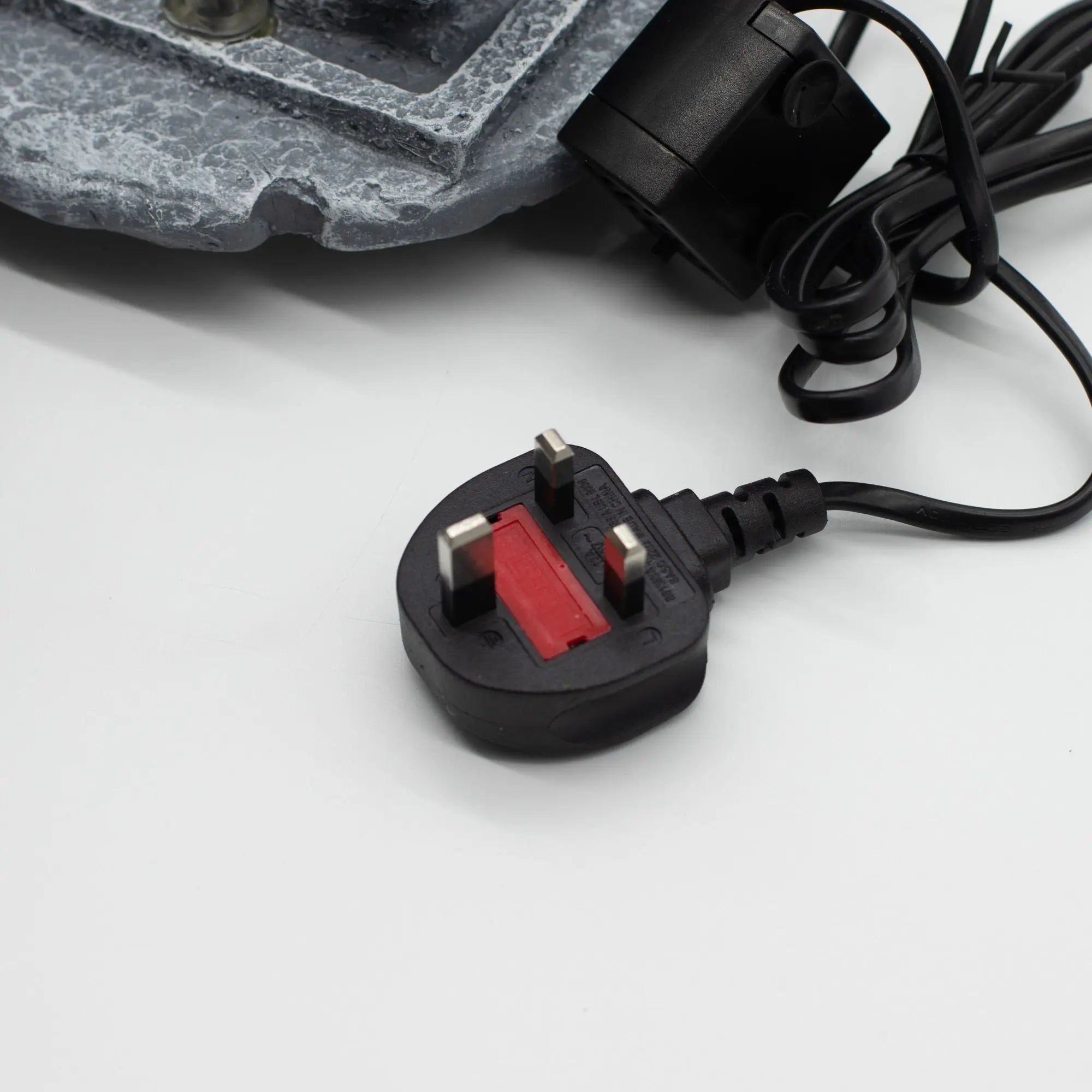 Close-up of a UK plug connected to the Tabletop Water Feature - 19x19x28cm - Buddha, Lotus Flower Cascading Potsx, showing part of the water feature base and electrical cord.