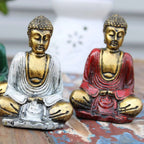 Two Gold Mini Buddha (Assorted Colours) statues sitting cross-legged in meditative poses with detailed gold faces and textured robes in white and red colors, displayed on a rustic wooden surface.