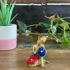 Brass Buddha Figure - Thinking - 6.5 cm seated in a contemplative pose holding colorful cushions, placed on a wooden surface with green plants in the background.
