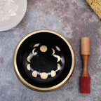 Large Moon Phase Singing Bowl Set- Black 14cm featuring gold moon phase designs inside a black bowl with a wooden striker on a textured surface.