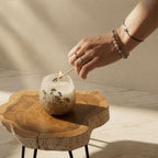 Hand lighting a Small Glass Jar Candles - Wildlight Woods on a natural wood slice table, showcasing delicate floral designs and a cozy, earthy ambiance.