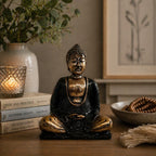 Black & Gold Buddha - Medium resin statue seated in meditation pose on a wooden surface, surrounded by books, a lit candle, and decorative elements in a calm, cozy room setting.