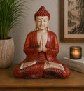 Hand Carved Buddha Statue - 40cm Welcome seated in a meditative pose with hands in prayer, featuring a red and cream finish, placed on a wooden surface beside a plant and candle.
