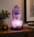 Buddha Bust Lamp Lavender Crackle - Oval Stand glowing softly in lavender on a black oval base, adding serene Buddha decor ambiance to a wooden table with plants and candles nearby.