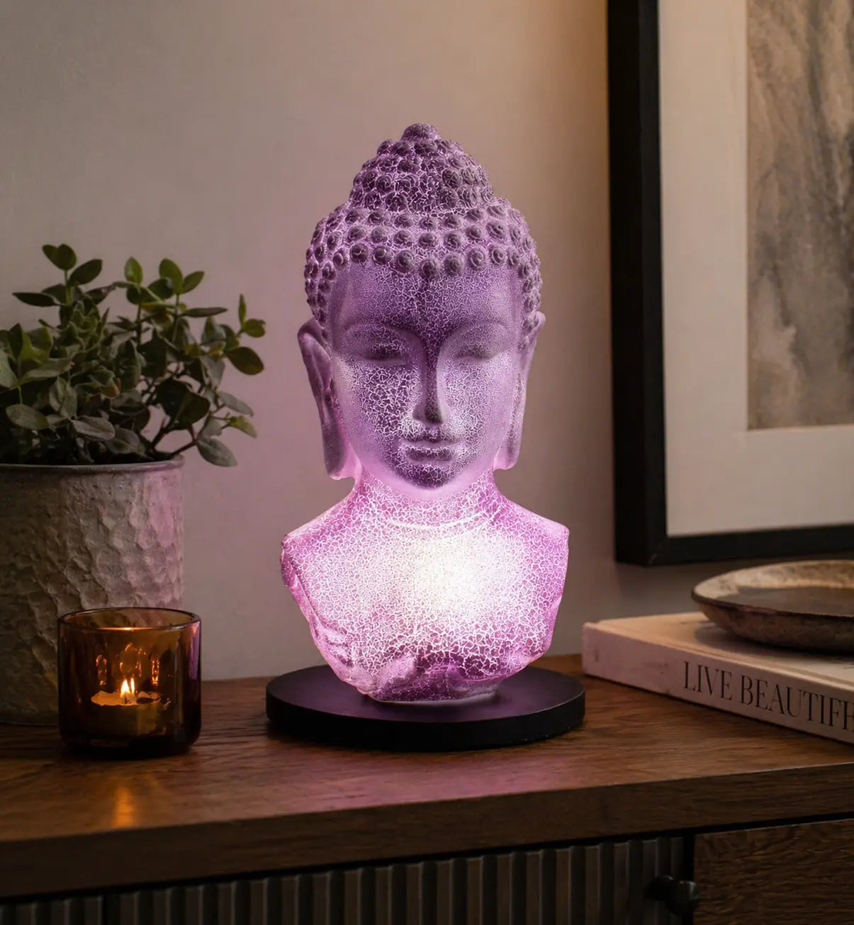 Buddha Bust Lamp Lavender Crackle - Oval Stand glowing softly in lavender light on a wooden surface, beside a candle, plant, and books, creating serene Buddha decor ambiance.