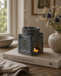 Square Buddha Burner - Dusty Blue sitting on a book with a lit candle inside, releasing smoke from the top, surrounded by soft natural light and a vase of flowers nearby.