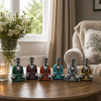 A row of six Silver Mini Buddha (Assorted Colours) statues in vibrant green, red, orange, turquoise, silver, and gold finishes displayed on a wooden table near a candle and flowers.