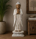Buddha Statue Standing - Whitewash - 1m Welcome displayed indoors on a wooden surface with a serene expression, hands in prayer, standing on a lotus base in rustic decor setting.