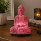 Meditation Buddha Lamp Cherry Crackle - Oval Stand glowing with a serene pink crackle finish, sitting on a wooden surface next to a potted plant and decorative candle.