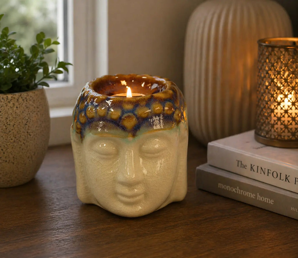 Buddha Oil Burner - Ivory & Mint ceramic oil burner in the shape of a serene Buddha head with a lit candle inside, displayed on a wooden surface next to books and decorative items.