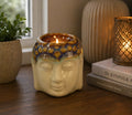 Buddha Oil Burner - Ivory & Mint ceramic oil burner in the shape of a serene Buddha head with a lit candle inside, displayed on a wooden surface next to books and decorative items.